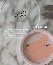 Ohra Self Made Eye Mask in its case, reusable, 100% medical-grade silicone, hypoallergenic and gentle on delicate skin. An open round makeup compact on a white marble surface, showing two peach-colored silicone eye patches inside a pale pink case. The lid is clear and branded 'ohra'.