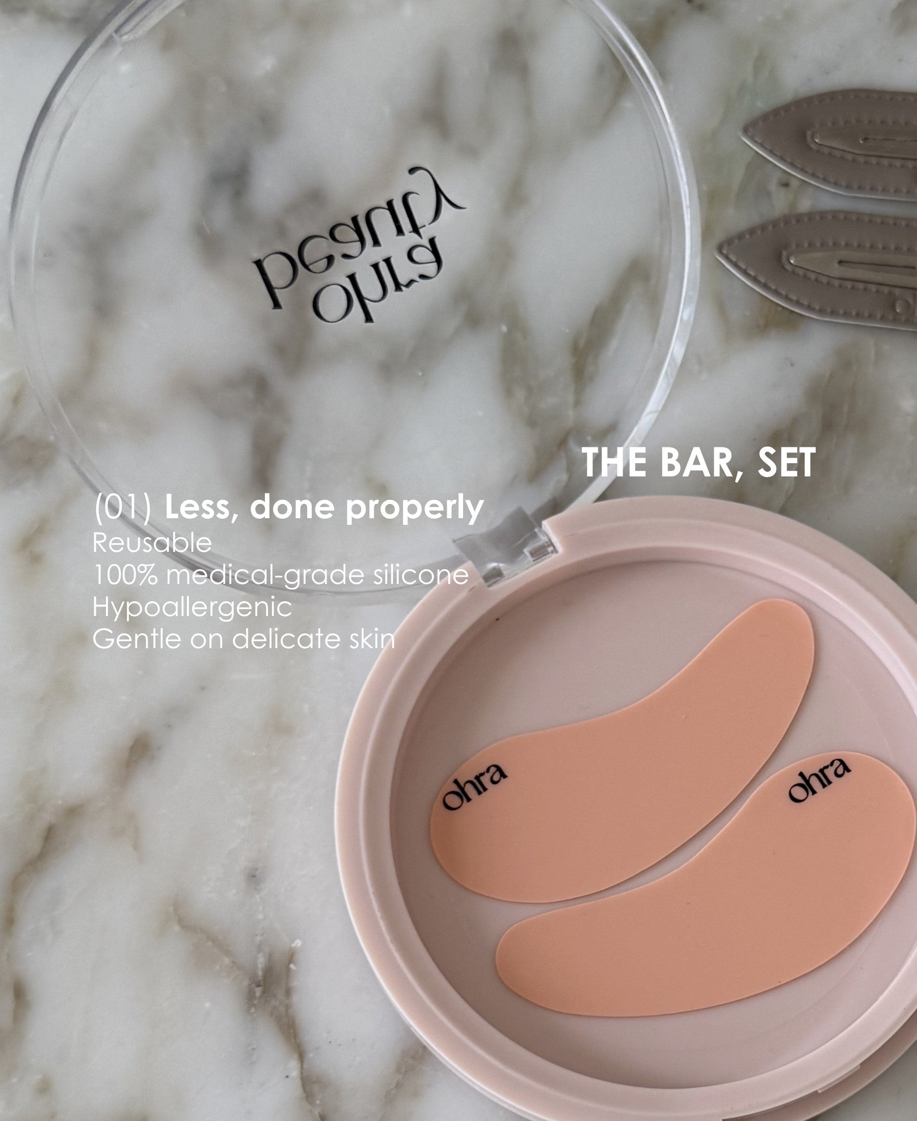 Ohra Self Made Eye Mask in its case, reusable, 100% medical-grade silicone, hypoallergenic and gentle on delicate skin. An open round makeup compact on a white marble surface, showing two peach-colored silicone eye patches inside a pale pink case. The lid is clear and branded 'ohra'.