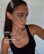 Woman wearing Ohra Untouched Hair Clips in her hair, styled with makeup during her application. A woman with long dark brown hair wearing a silver feather hair clip, shown in a close-up portrait against a white brick wall. Text on the image reads 'OHRA UNTOUCHED HAIR CLIPS'.