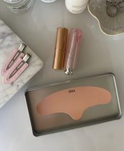 Flat lay of the Ohra Self Made Eye Mask storage tin and Untouched Hair Clips in Blush, styled with makeup on a marble surface