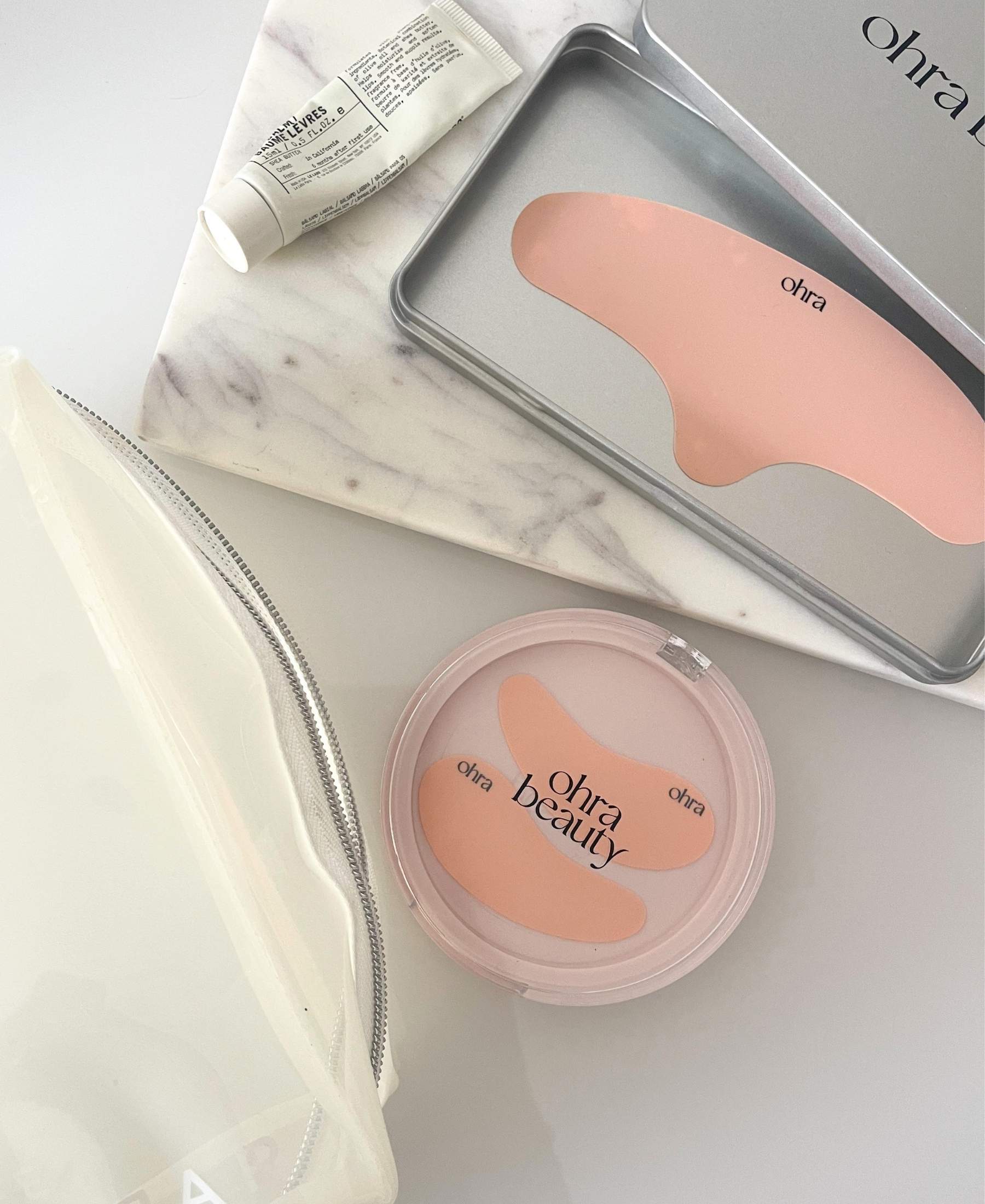 Flat lay of the Self Made Eye Mask and Lifted Forehead Mask cases styled with skincare and a clear makeup pouch on marble
